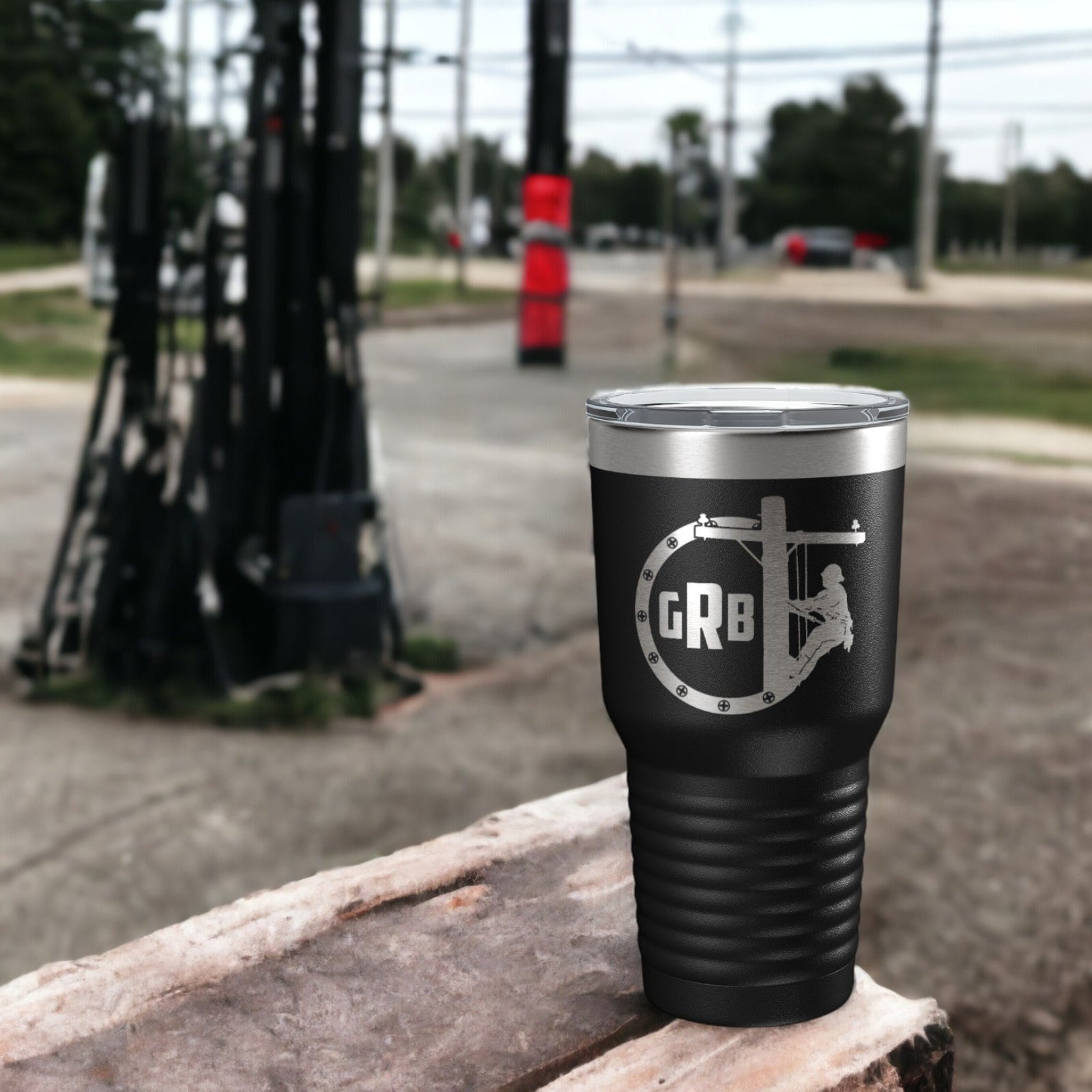A black stainless steel tumbler with a personalized monogram design, placed on a wooden surface with a background featuring utility poles and a red traffic cone.