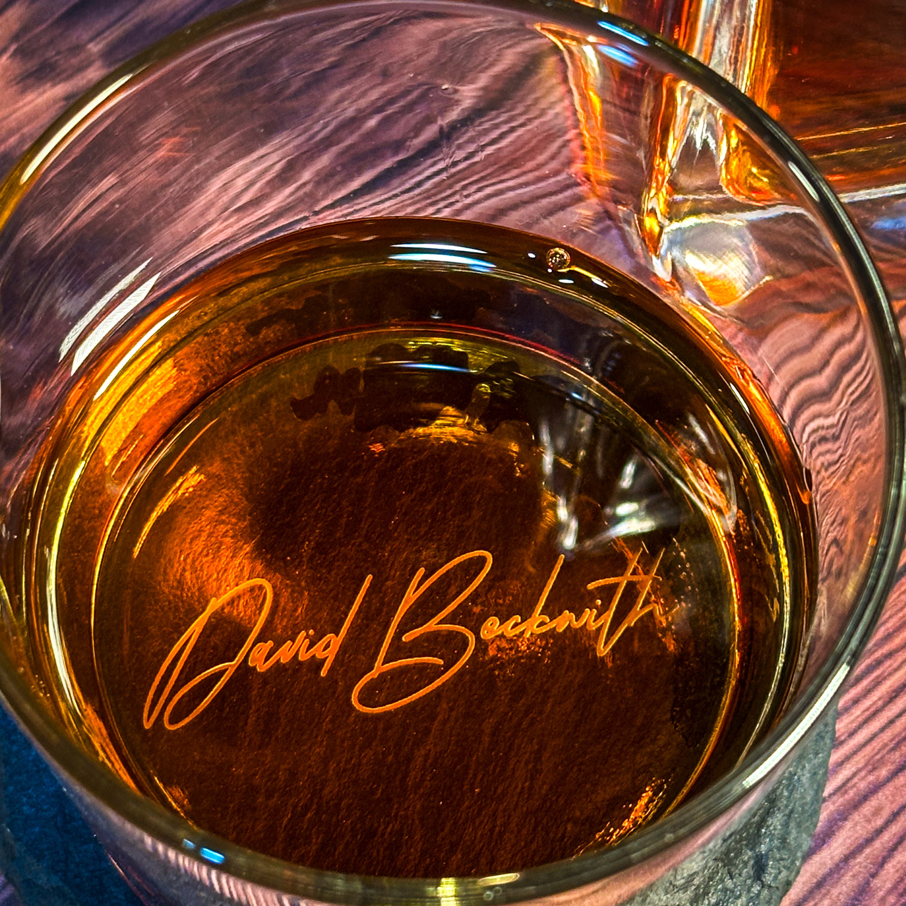 A close-up of an engraved whiskey glass with the name 'David Beckham' sandblasted into the glass, filled with whiskey.